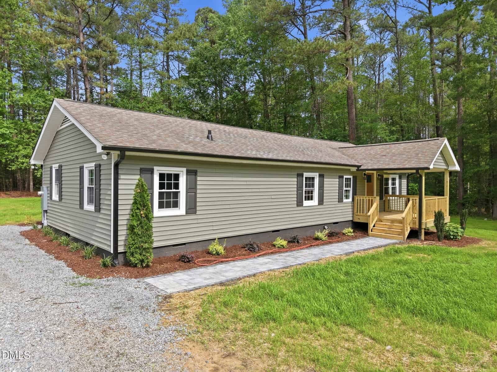 A single-story gray house with white trim, a shingled roof, black shutters, and a small covered front porch, surrounded by a well-maintained lawn and trees in the background.