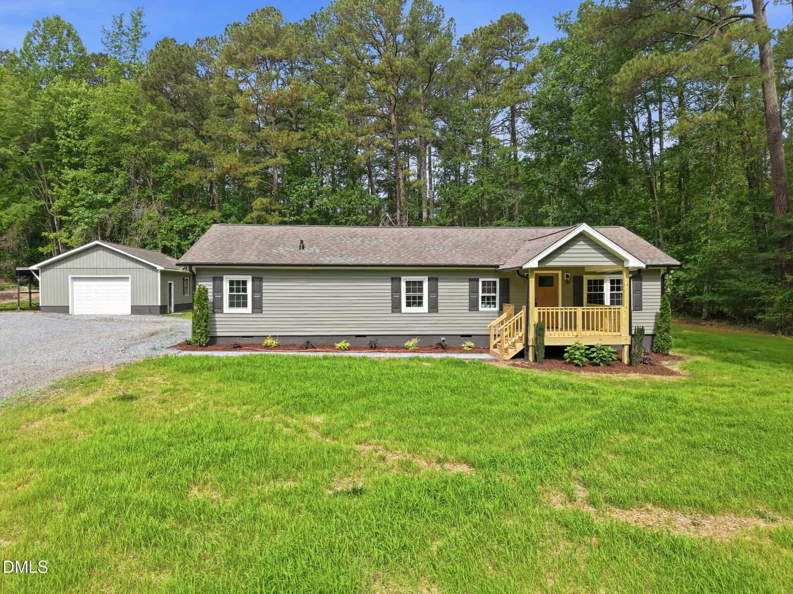 A single-story gray house with a small covered porch, surrounded by green grass and trees, with a detached garage and gravel driveway to the left.
