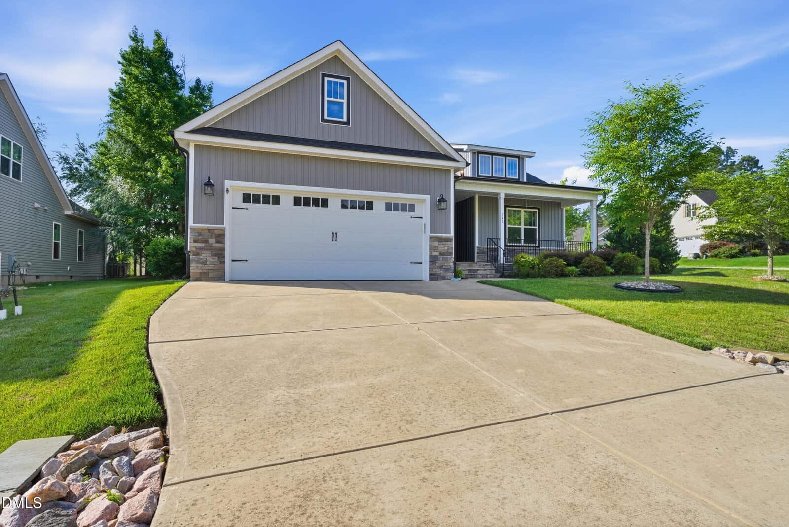 A modern suburban house with gray siding, white trim, and a double garage. The driveway is wide and clean, and the front yard is neatly landscaped with green grass, shrubs, and small trees.