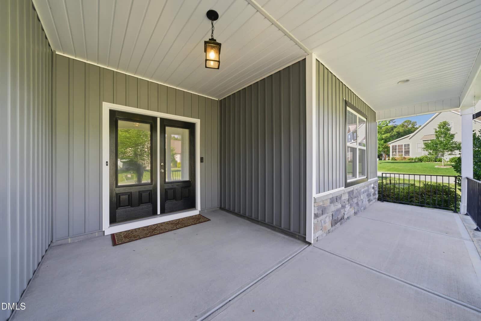 Covered front porch with double black doors, gray vertical siding, stone accents, a small hanging light, and a welcome mat, overlooking a concrete walkway and a green yard in a suburban neighborhood.