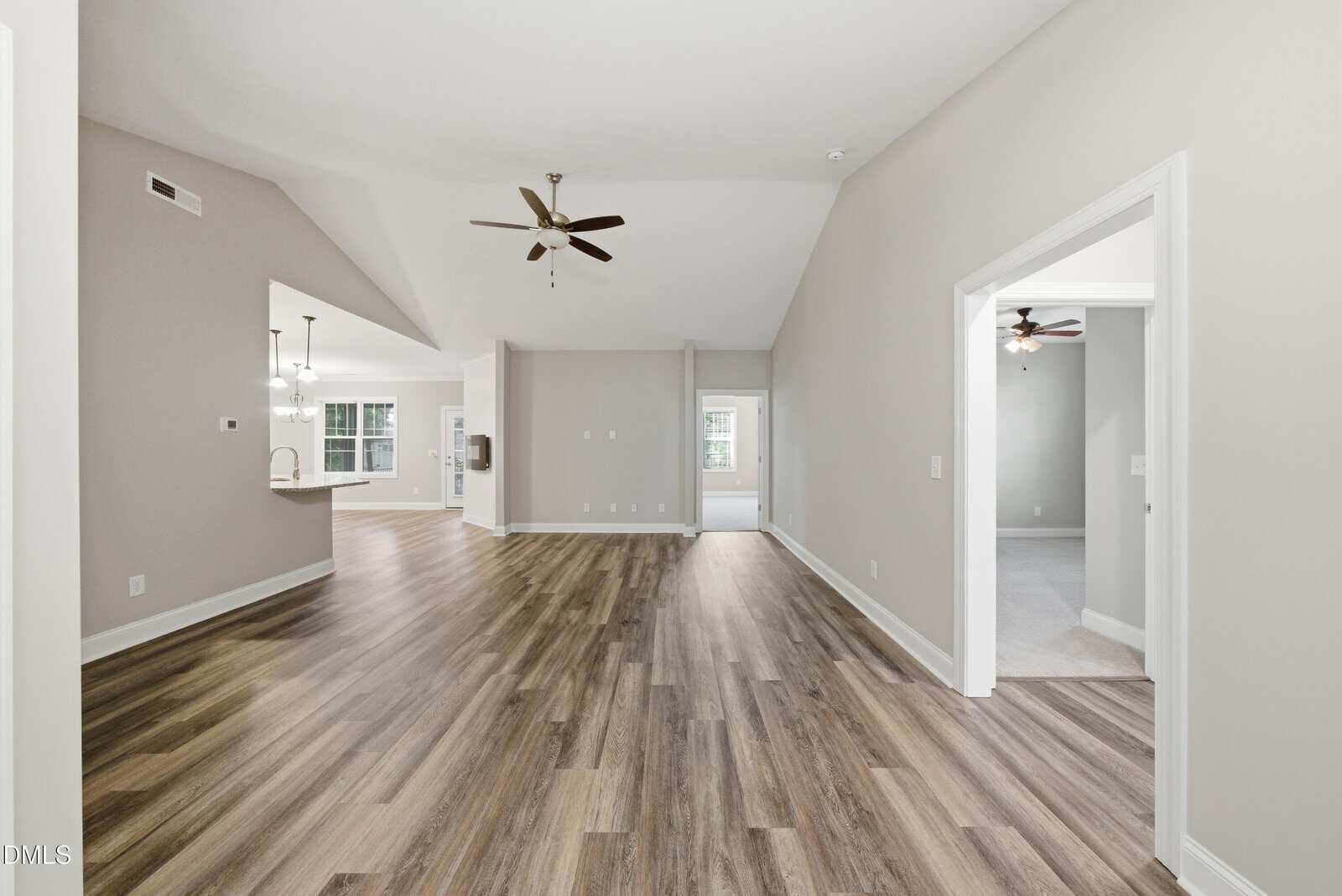 Spacious, modern living area with wood-style flooring, neutral walls, ceiling fans, and open access to a kitchen, hallway, and two carpeted bedrooms with large windows.