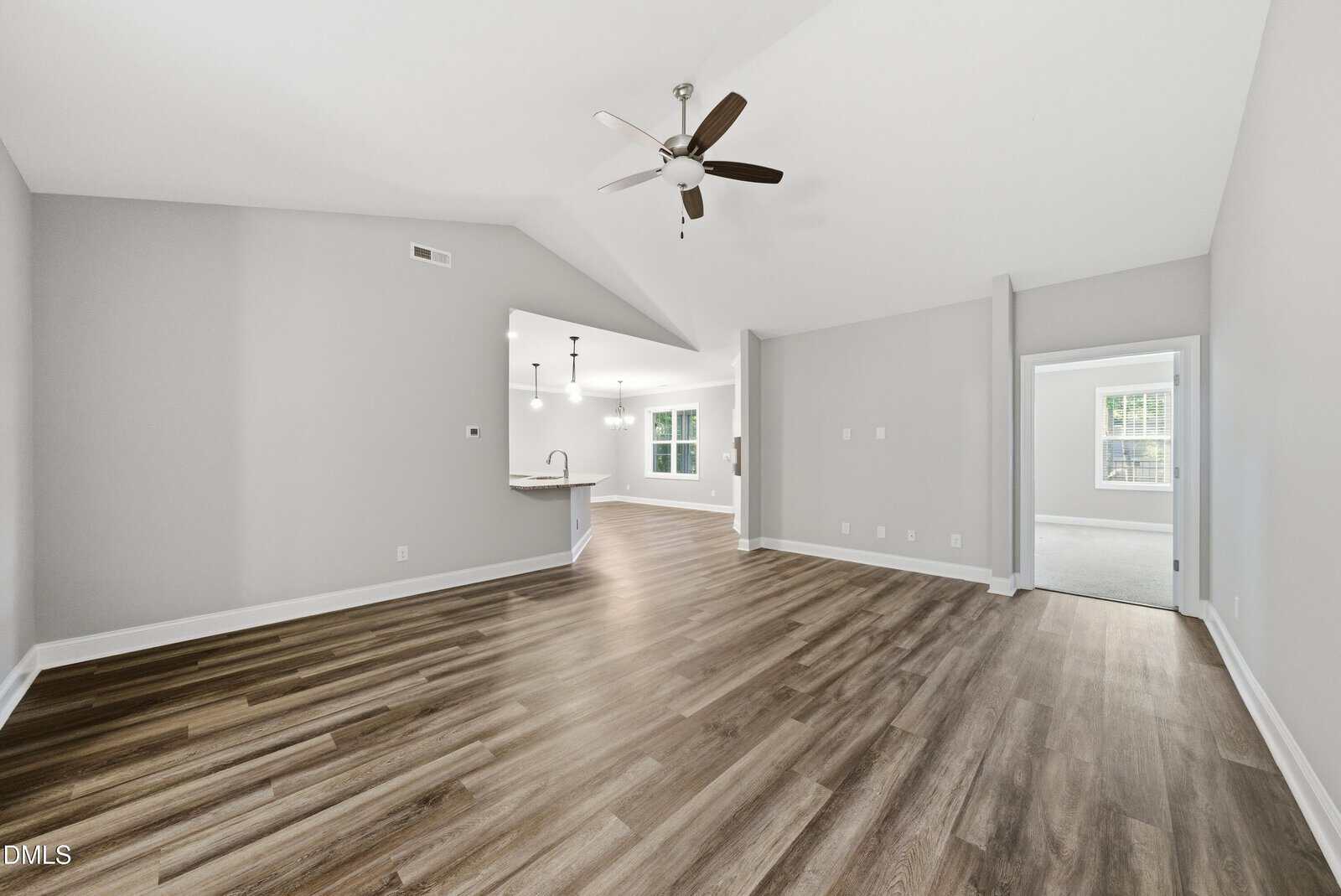 Spacious open-concept living area with wood-style flooring, neutral walls, a ceiling fan, and view into a modern kitchen with pendant lights. Large windows provide natural light; a doorway leads to a carpeted room.