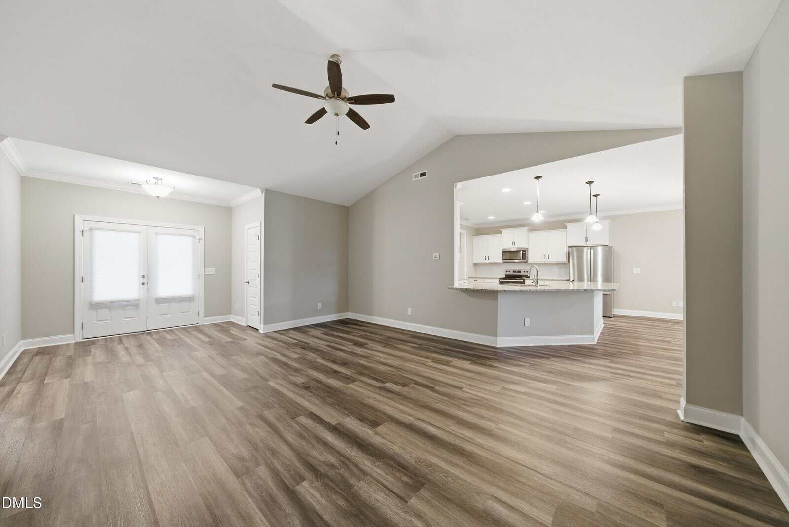 Spacious living room with wood-look flooring, neutral walls, ceiling fan, and view into modern kitchen with white cabinets and stainless steel appliances. Double doors lead outside.