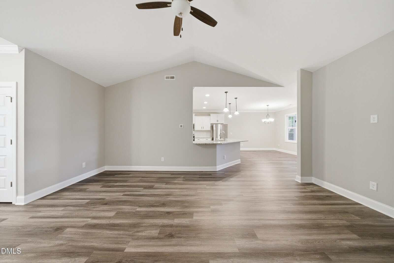 Spacious, empty room with light gray walls, wood-look flooring, ceiling fan, and an open view into a modern kitchen and dining area with pendant lights and large windows.