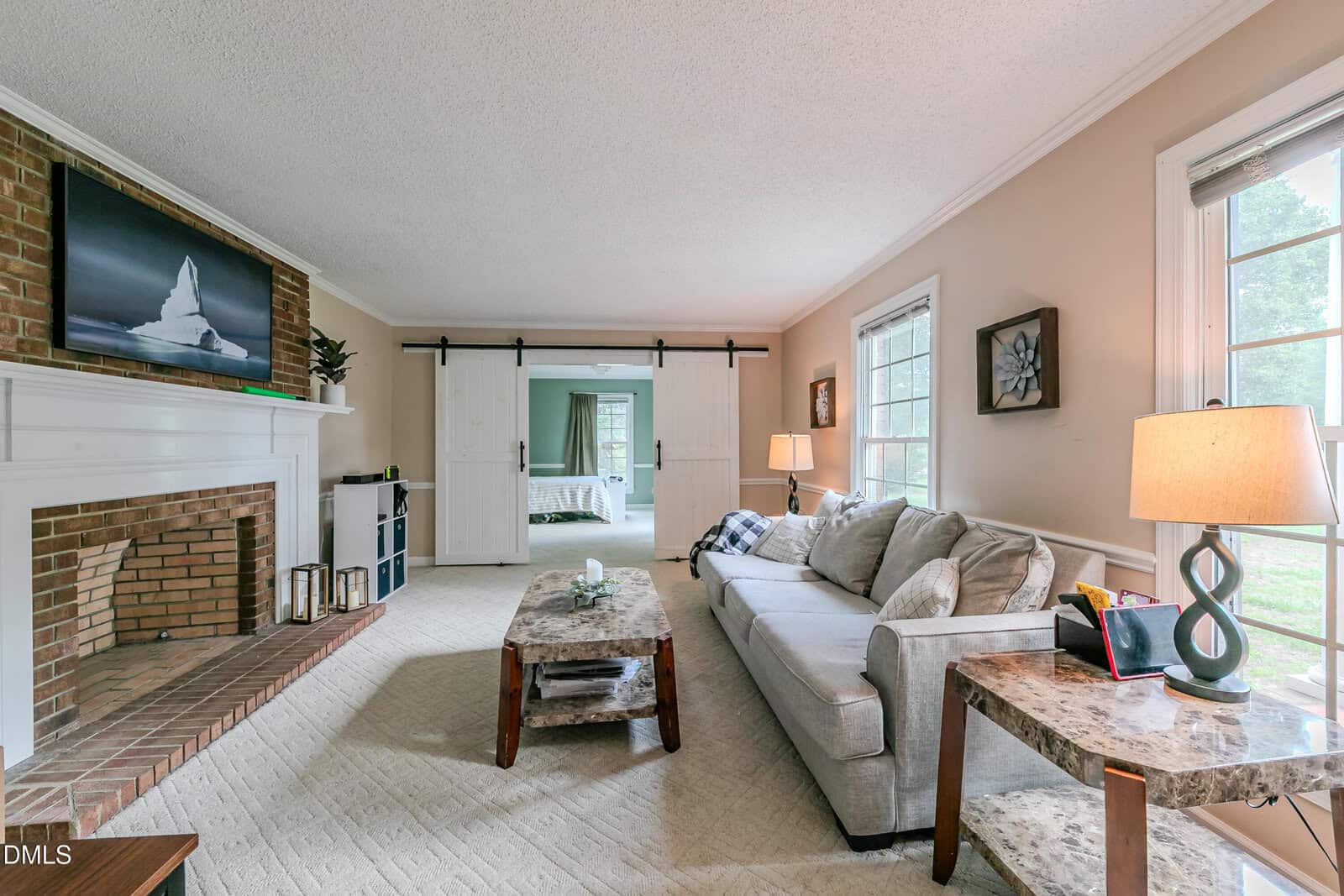 Bright living room with a brick fireplace, a beige sofa, and marble-topped tables. Large windows let in natural light. Sliding barn doors open to a bedroom in the background. Decor includes artwork and a table lamp.