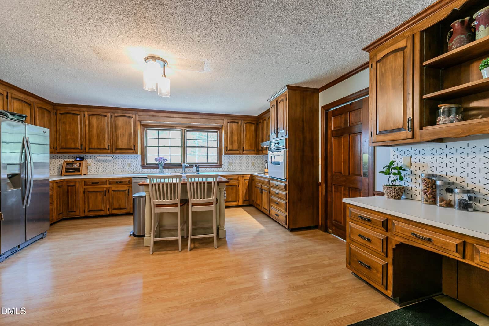 Spacious kitchen with wooden cabinets, stainless steel appliances, a center island with two chairs, wood flooring, a window over the sink, and a built-in desk area with plants and decorative tiles.