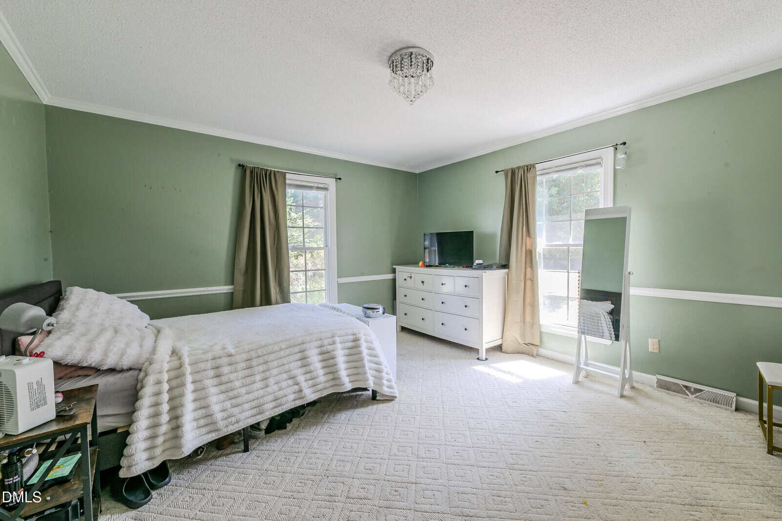 A bedroom with green walls, a bed with white bedding, a dresser with a TV, a full-length mirror, two windows with curtains, and a small chandelier on the ceiling.