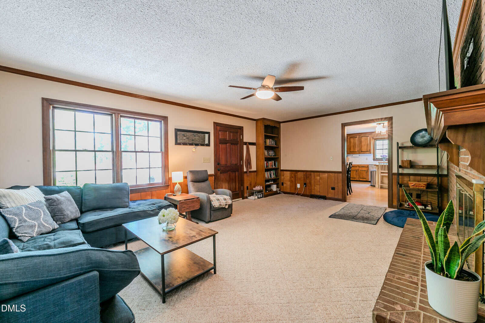 Spacious living room with beige carpet, blue sectional sofa, coffee table, built-in shelves, brick fireplace, ceiling fan, and large windows letting in natural light. Kitchen visible through doorway.