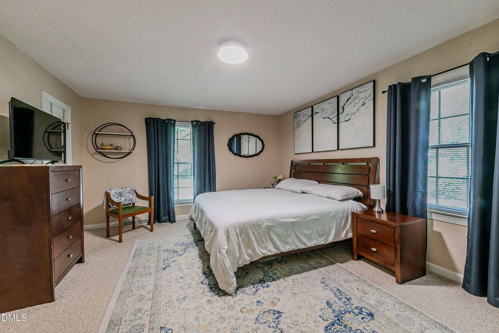 A cozy bedroom with a large bed, dark wood furniture, dark curtains on windows, a TV on a dresser, a chair, a patterned rug, and abstract wall art above the headboard. Natural light fills the room.