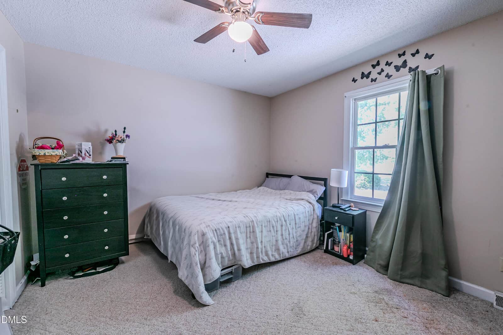 A simple bedroom with a bed, black dresser, nightstand, lamp, and green curtain. There are butterfly wall decals above the window, and sunlight comes through the window onto the beige carpeted floor.