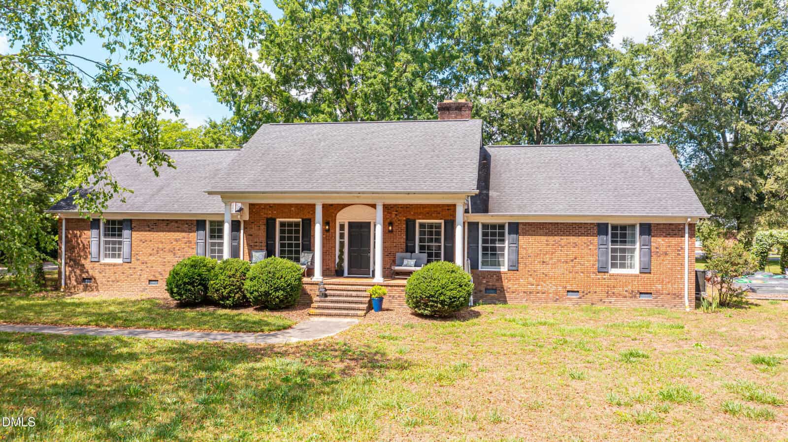 A single-story brick house at 8008 NC 39 Hwy in Zebulon features a gray roof, white columns, and black window shutters, surrounded by trees and shrubs under a bright blue sky.