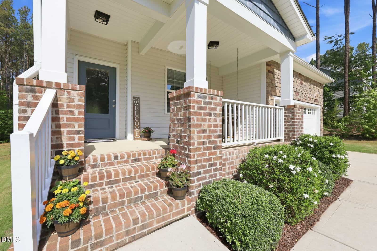 A front porch with brick steps, potted flowers, a "Welcome" sign, white railings, and a light blue front door. Bushes and greenery line the concrete walkway beside the house.