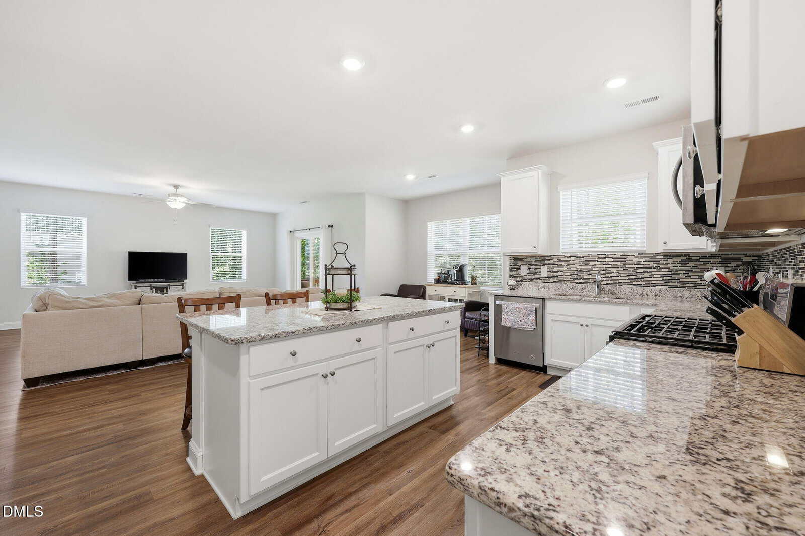Bright, modern kitchen with white cabinets, granite countertops, a large island, stainless steel appliances, and wood floors. The kitchen opens to a living area with a beige sectional sofa and a TV. Large windows provide ample natural light.