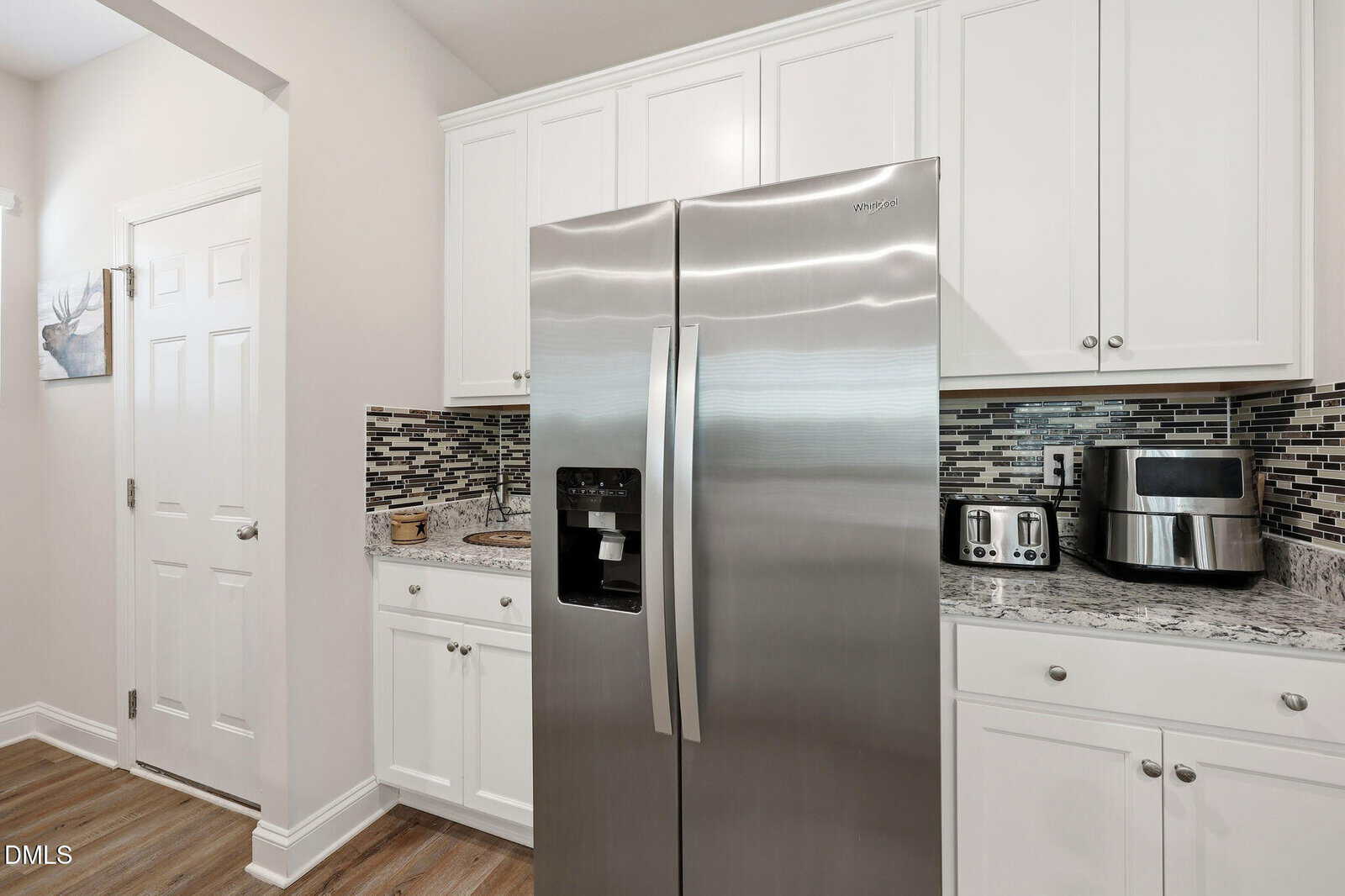 Modern kitchen with white cabinets, stainless steel refrigerator, toaster, and air fryer on granite countertops, and a mosaic tile backsplash. Light wood floor and a closed white door are also visible.
