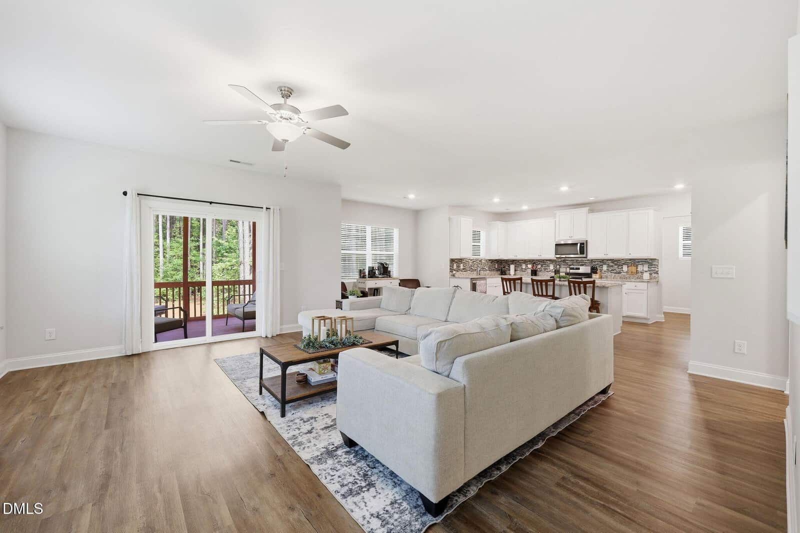 Bright, open-concept living room and kitchen with light walls, wood floors, a large beige sectional sofa, modern coffee table, dining area, and sliding doors leading to a screened porch with outdoor seating.