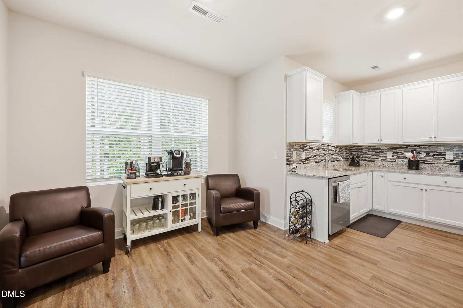 Bright kitchen and sitting area with two brown armchairs, a coffee station on a white sideboard, wood-style flooring, large window with blinds, and white cabinets with a tiled backsplash.