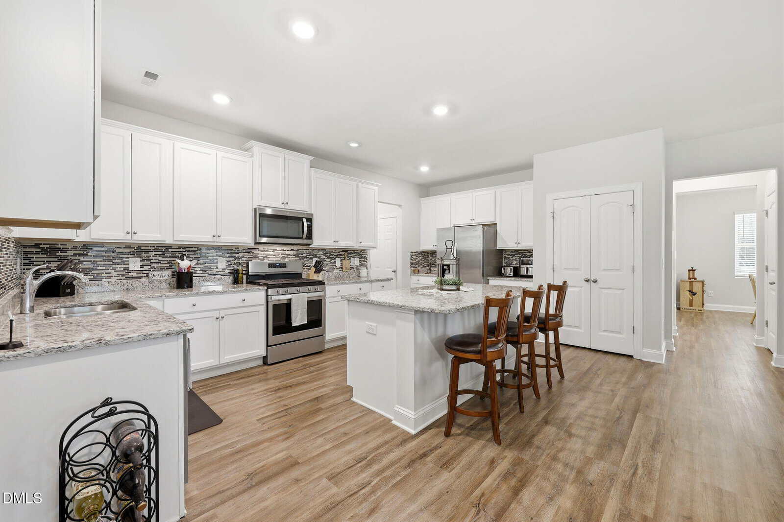 Modern kitchen with white cabinets, stainless steel appliances, an island with two wooden chairs, granite countertops, tile backsplash, and wood-style flooring. A hallway and a partially visible room are in the background.