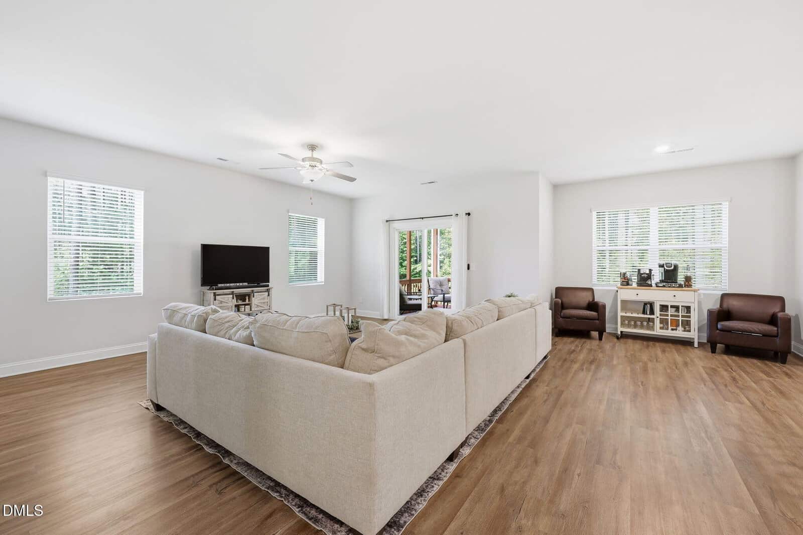 Modern living room with a large beige sectional sofa, TV, two leather chairs, a console with decor, wood flooring, white walls, and windows letting in natural light. Glass doors lead to an outdoor area with greenery.