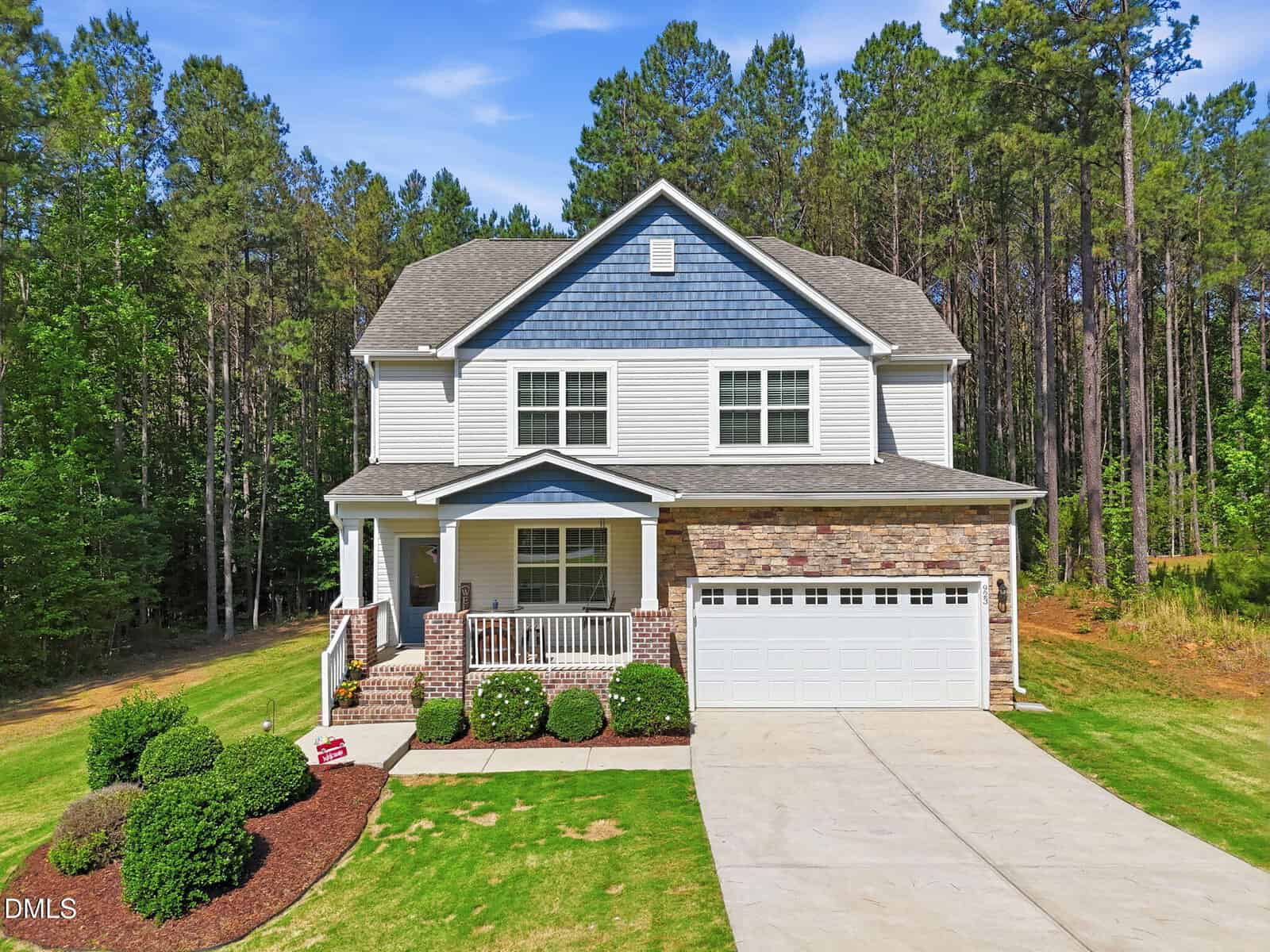 Charming two-story suburban house at 923 Weatherby Lane, Creedmoor, with blue and white siding, a front porch, attached two-car garage, and landscaped yard surrounded by tall trees and greenery under a clear sky.