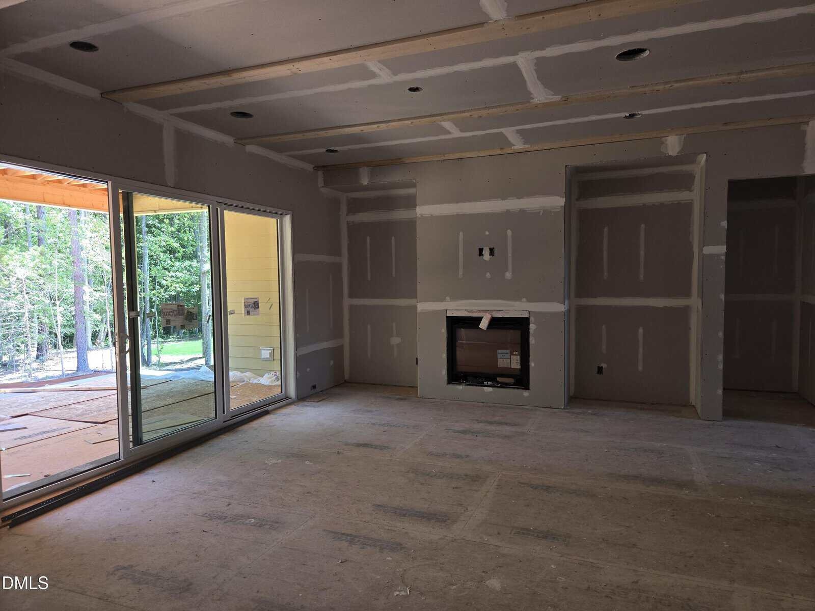A partially finished room with drywall installed, exposed seams, and an unfinished fireplace. Large glass doors open to a covered patio with a view of trees outside. No flooring or paint is present yet.