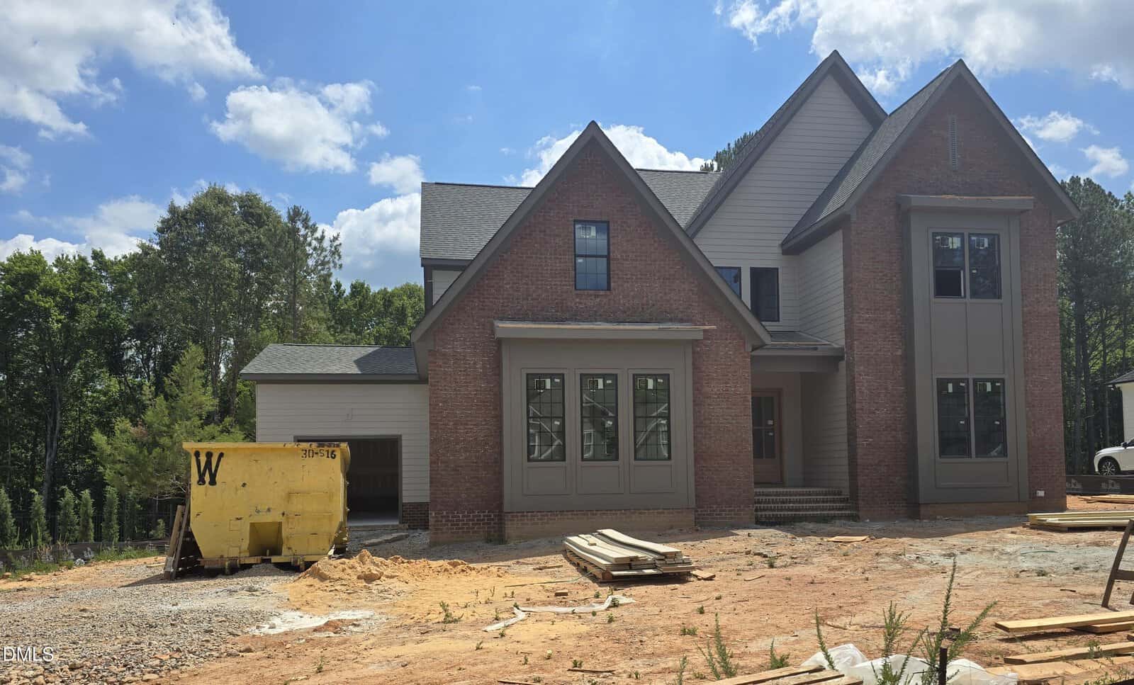 A two-story house under construction with red brick and gray siding, featuring large front windows. Construction materials and a yellow dumpster are in the dirt yard under a partly cloudy sky. Trees are visible in the background.