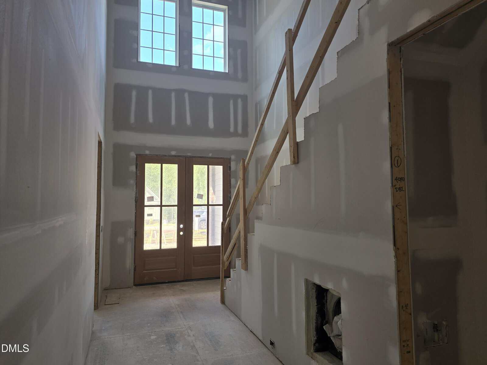 A hallway in a house under construction with unfinished drywall, a staircase with a temporary wooden railing, double front doors with glass panes, and large windows above letting in natural light.