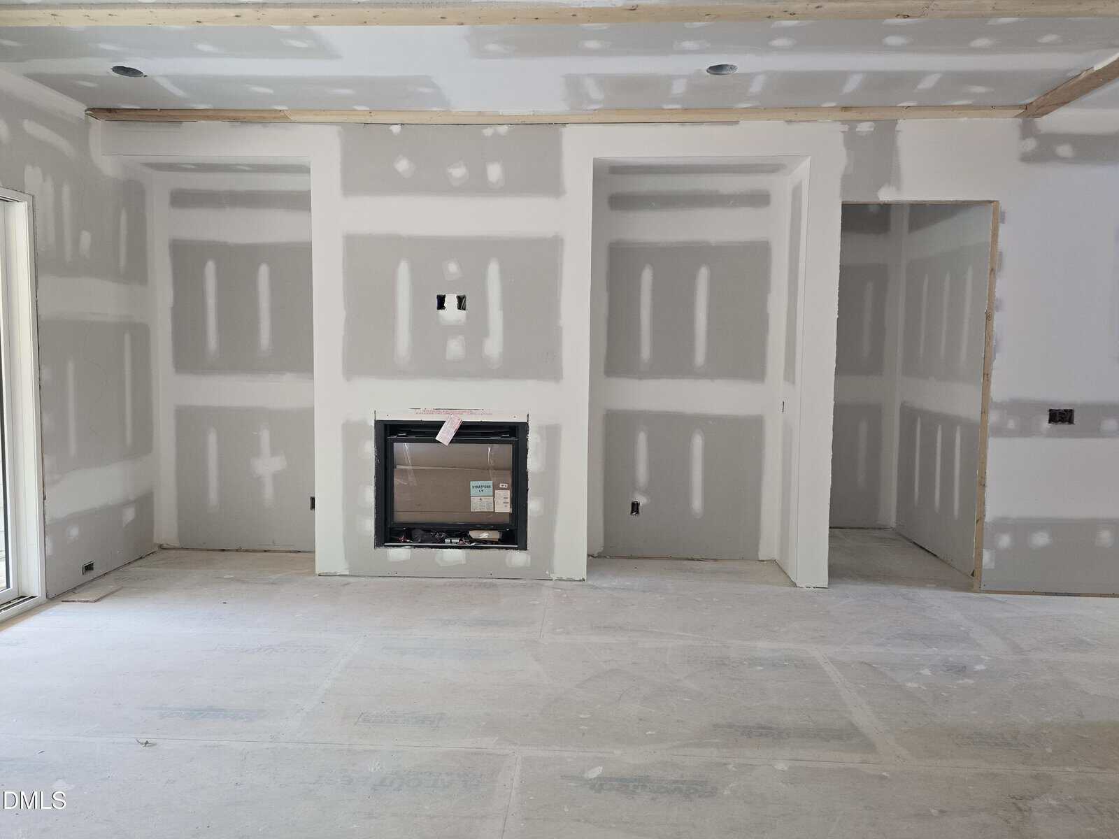 Unfinished living room with drywall installed, exposing taped seams and screw holes. There is a fireplace in the center of the wall and an open doorway to the right. The floor is covered for protection during construction.