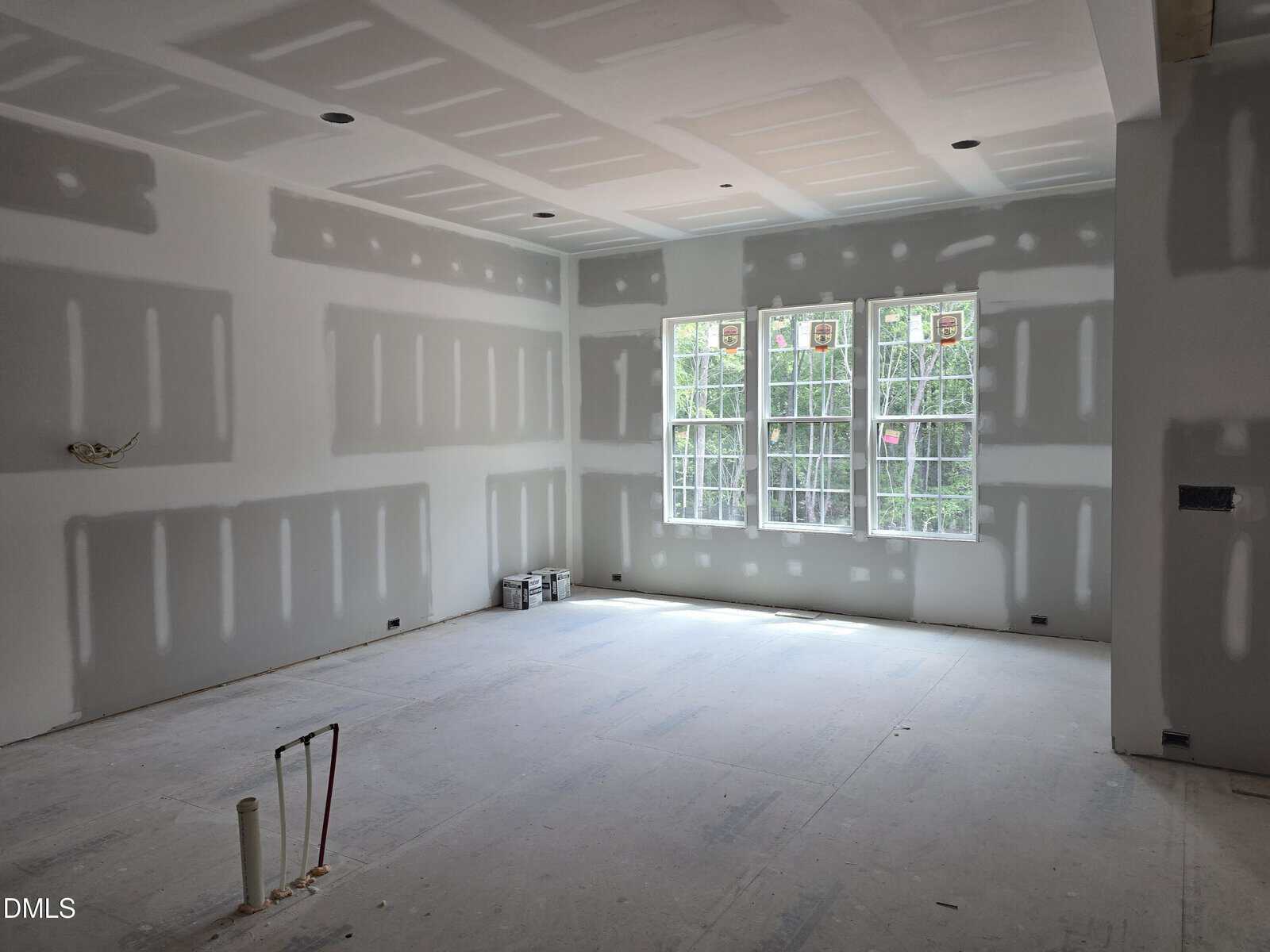 A room under construction with unfinished drywall, exposed joints, and no flooring. Sunlight streams in through three large windows. Construction materials are stacked against a wall.