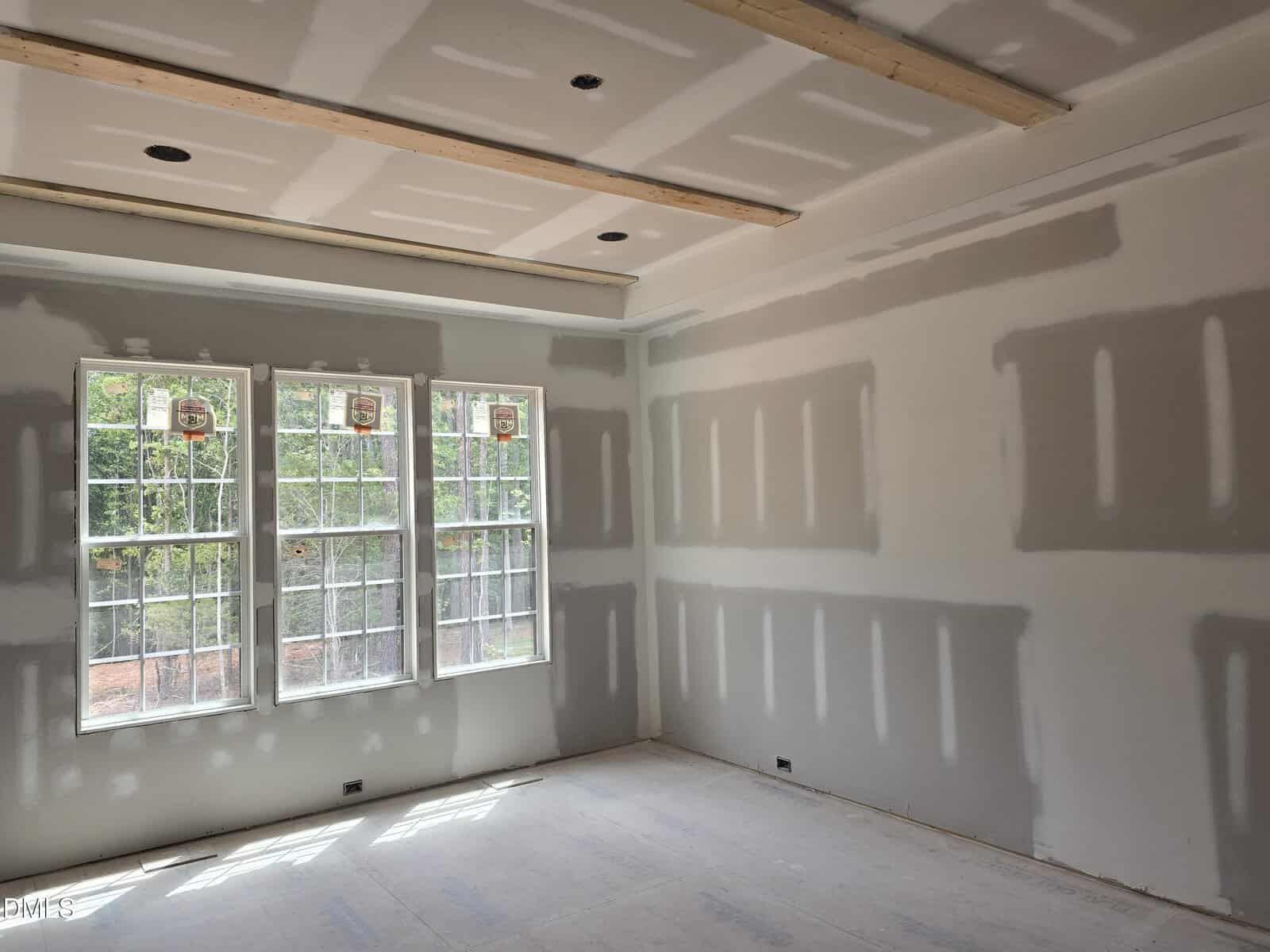 A room under construction with unfinished drywall, exposed seams, and three large windows letting in sunlight. The floor and ceiling are unfinished, and the walls show visible drywall tape and compound.