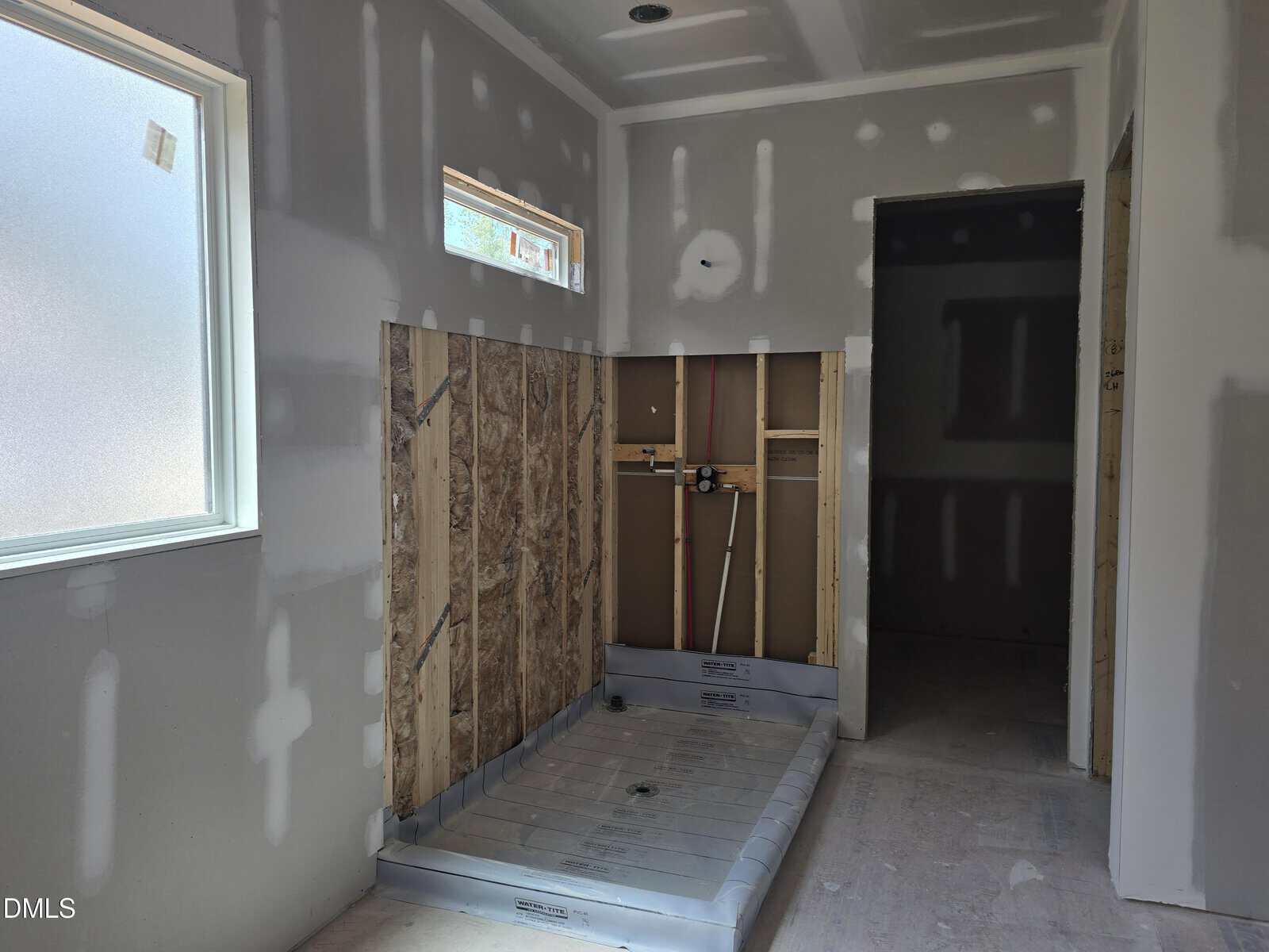 A bathroom under construction with unfinished drywall, exposed plumbing, a metal shower pan, and two windows providing natural light.