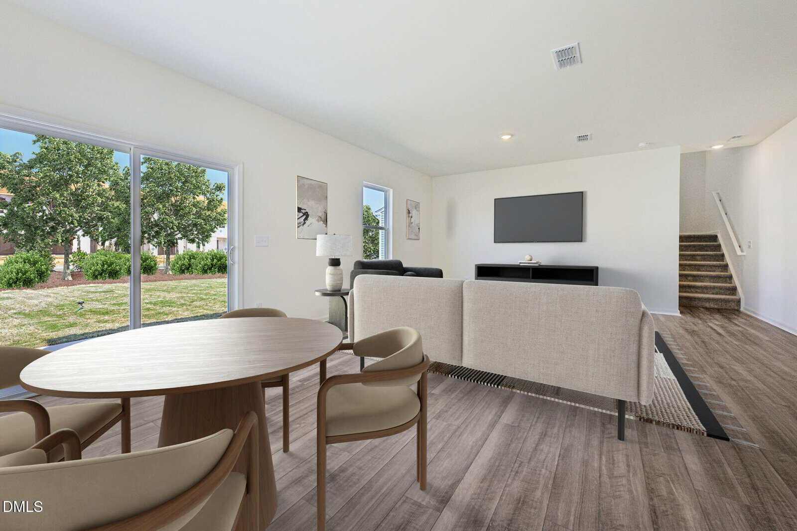 A bright, modern living room with light wood flooring, a beige sofa, round dining table with four chairs, TV on the wall, large windows, and a sliding glass door overlooking a green backyard. Stairs lead upstairs on the right.