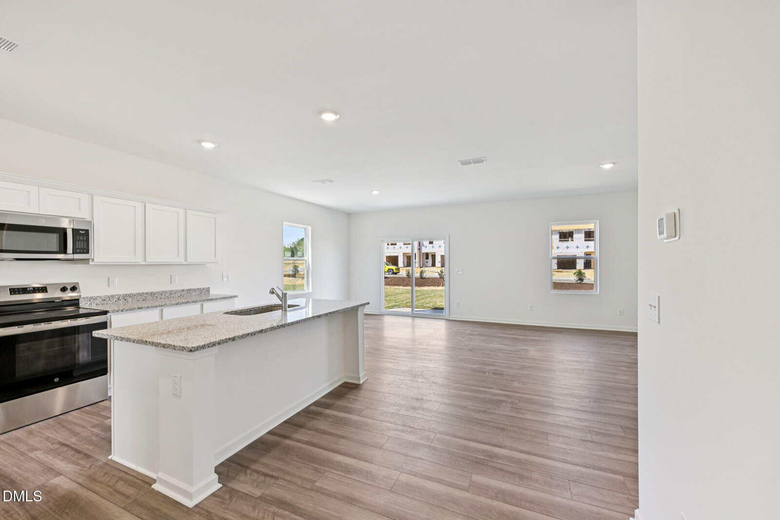 Modern open-concept kitchen and living area with white cabinets, stainless steel appliances, granite countertops, wood flooring, and large sliding glass doors leading to a grassy outdoor area with buildings in the background.