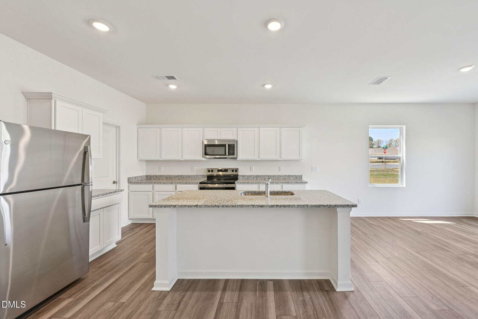 Modern kitchen with stainless steel appliances, white cabinets, a large island with sink, granite countertops, and wood-look flooring. Bright and spacious with ample natural light from a window.