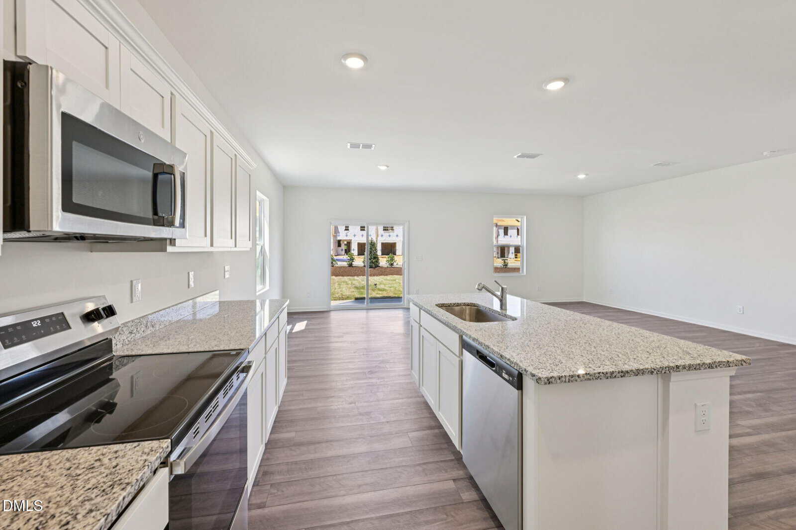 Modern kitchen with white cabinets, granite countertops, stainless steel appliances, an island with a sink, and open layout leading to a spacious living area with large windows and glass doors overlooking a grassy yard.