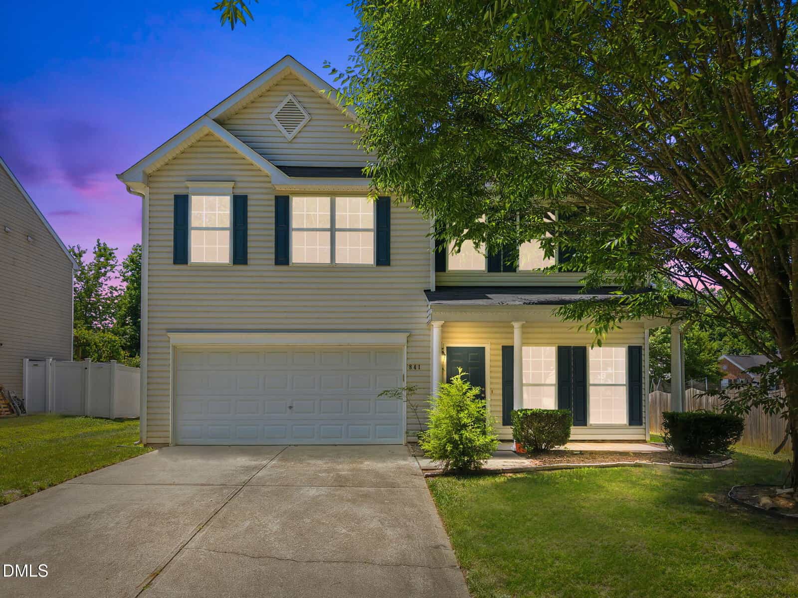 A two-story beige house with dark shutters at 841 Stackhurst Way, Wake Forest, featuring a double garage and covered front porch. Lit from inside, it sits on a green yard with a tree in front, photographed at dusk beneath a purple-blue sky.
