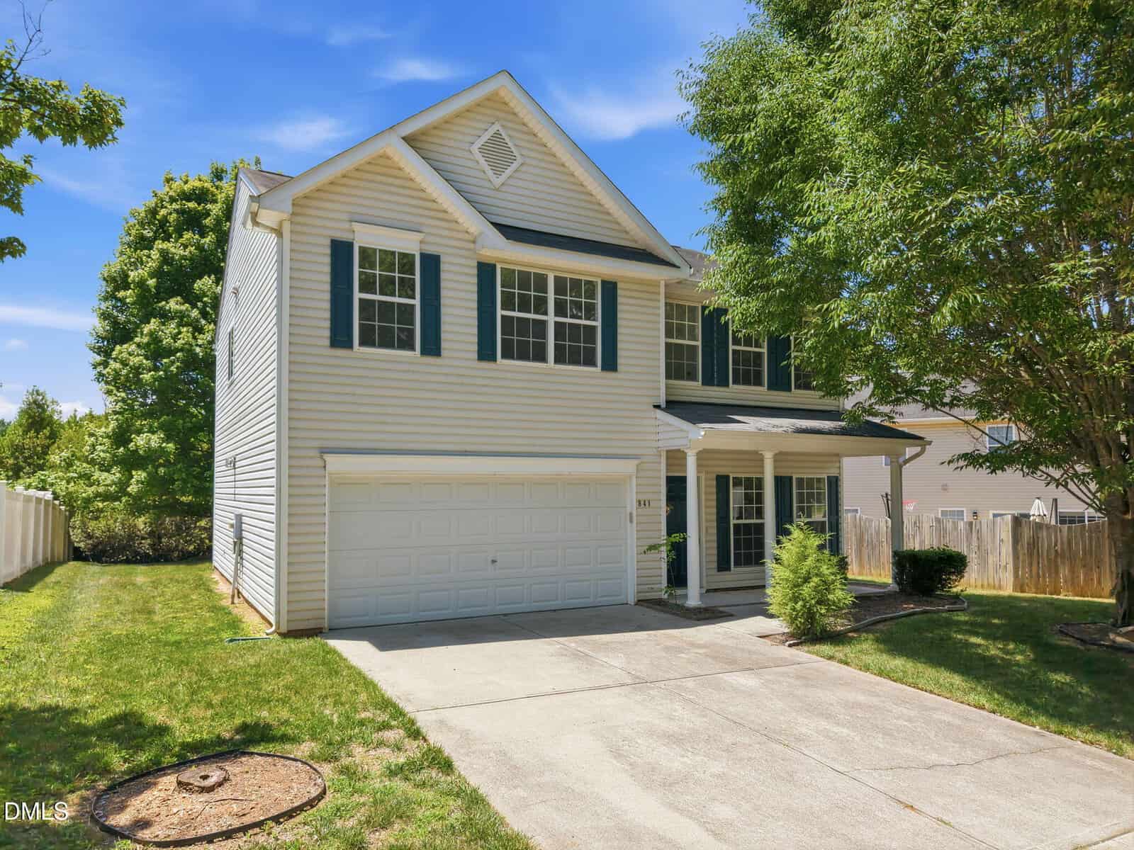 Two-story beige house with green shutters, a double garage, and a covered front porch. Concrete driveway, small front lawn, and trees surround the property under a blue sky.