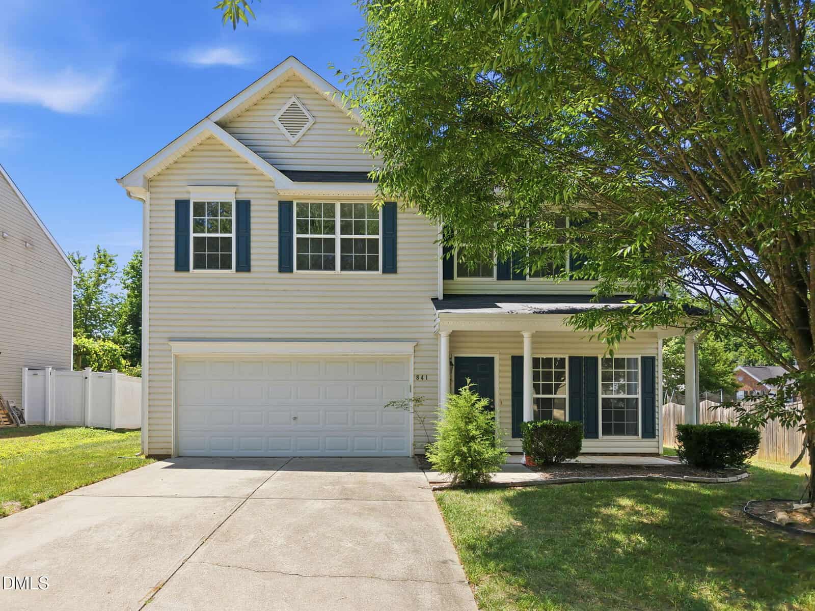 Two-story beige house with dark green shutters, a double garage, and a covered front porch. A large tree provides shade, and there is a driveway with a small landscaped area by the entry.
