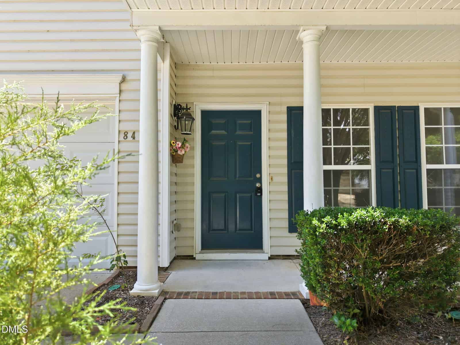 A front porch with white columns, a dark blue door, hanging lantern, flower basket, and large windows with blue shutters, surrounded by green bushes and plants. House number 84 is visible next to the door.