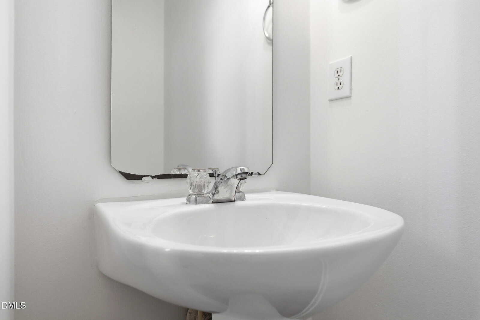 White pedestal sink with chrome faucet beneath a rectangular mirror on a white wall. Two electrical outlets are visible to the right of the mirror. The overall bathroom design is minimalistic.