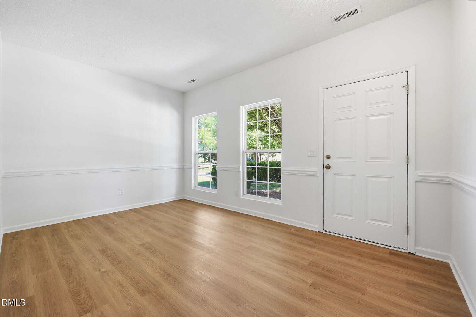 A bright, empty room with white walls, light wood flooring, two large windows, and a white front door, letting in natural light and showing greenery outside.