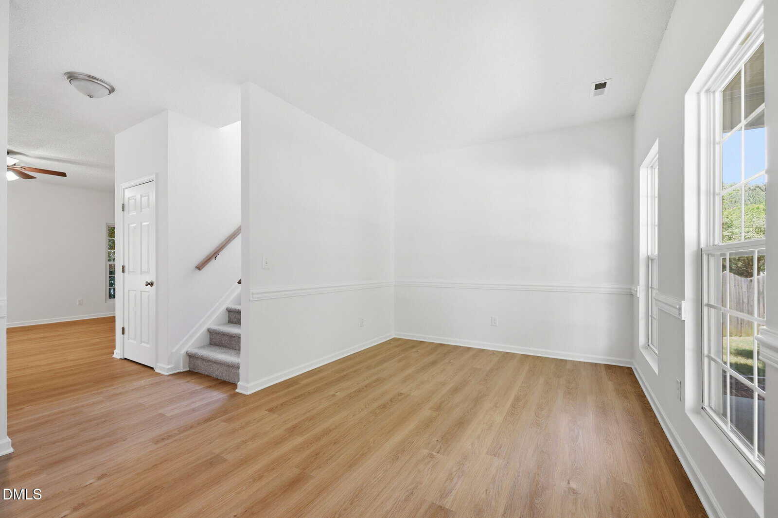Bright, empty room with white walls, large windows, light wood flooring, and a staircase with gray carpet leading upstairs. A doorway on the left opens to another room with hardwood floors.
