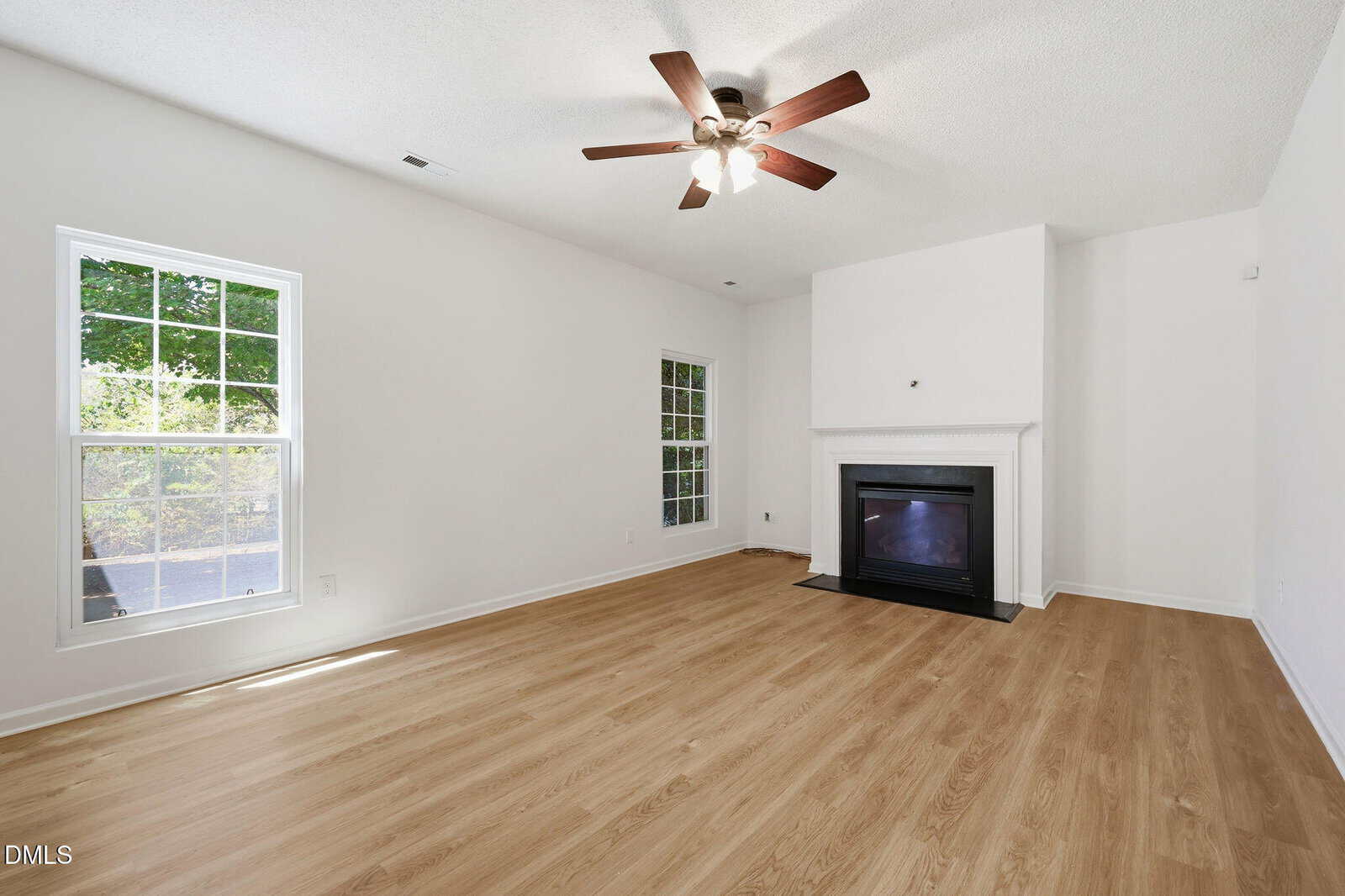 Bright living room with light wood flooring, white walls, two large windows, a ceiling fan with lights, and a black fireplace with a white mantel on the right wall.