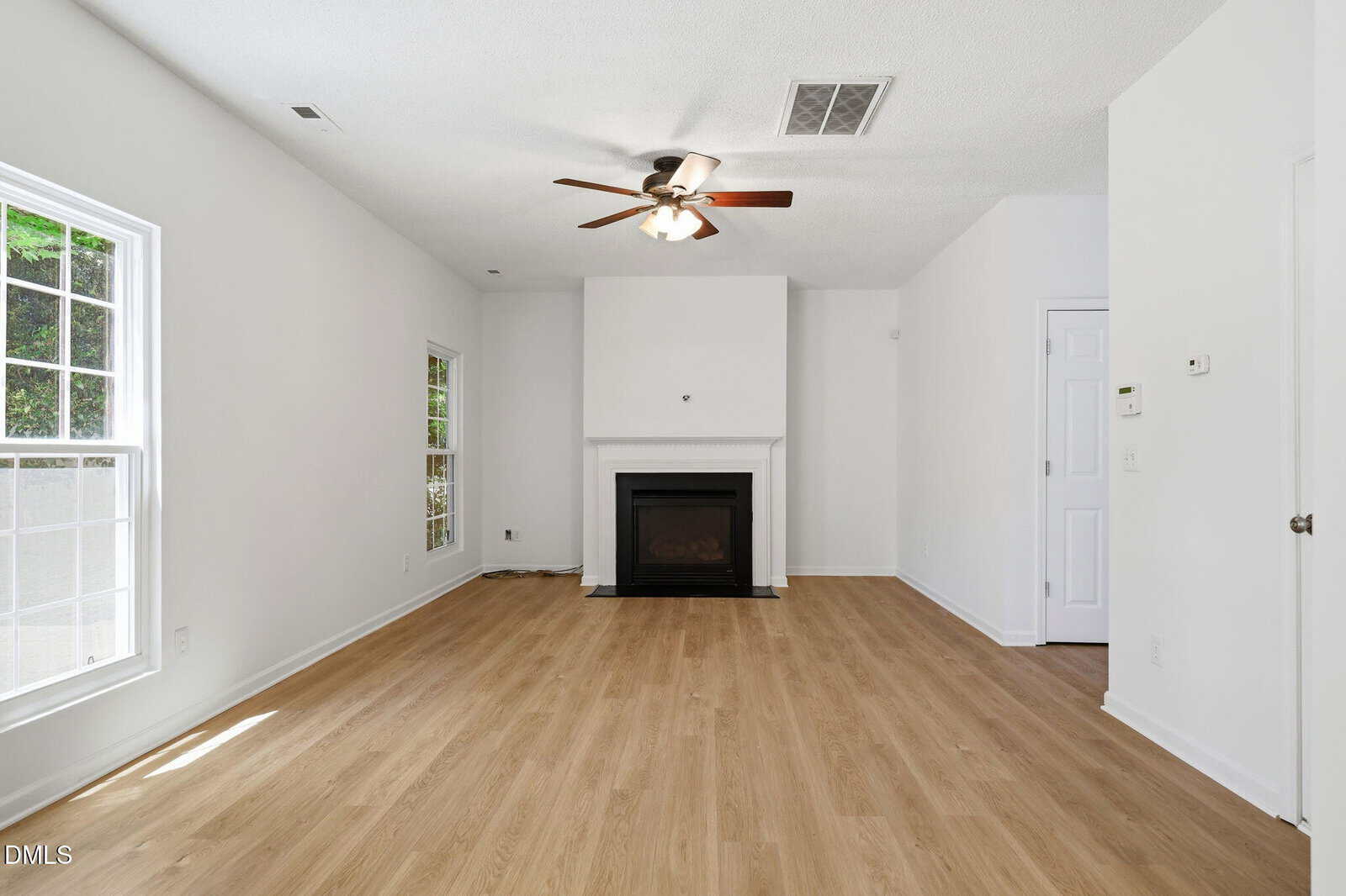 Bright living room with light wood floors, white walls, large windows, a ceiling fan, and a fireplace centered on the far wall. Natural light fills the room, giving it an open and inviting feel.