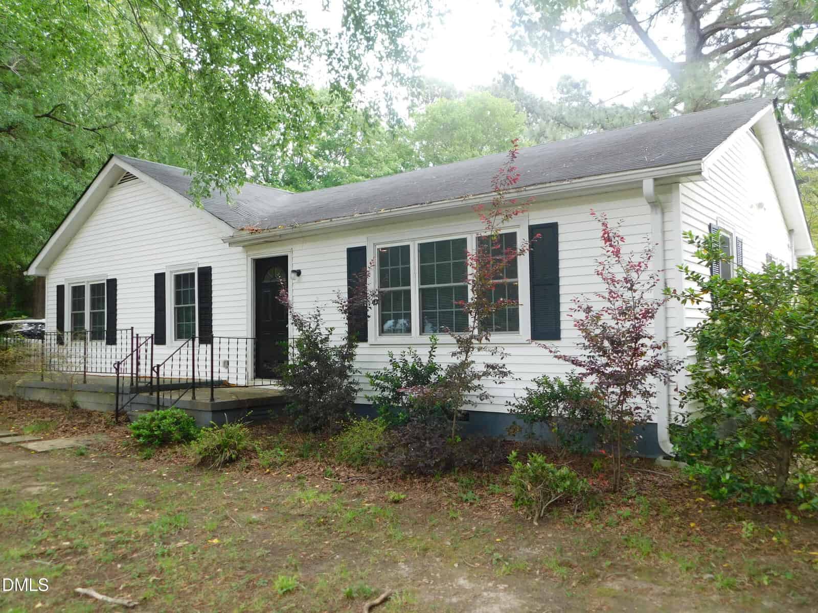 A single-story white house with black shutters and a dark roof, surrounded by small bushes and trees. There is a front porch with a metal railing and a slightly overgrown yard.