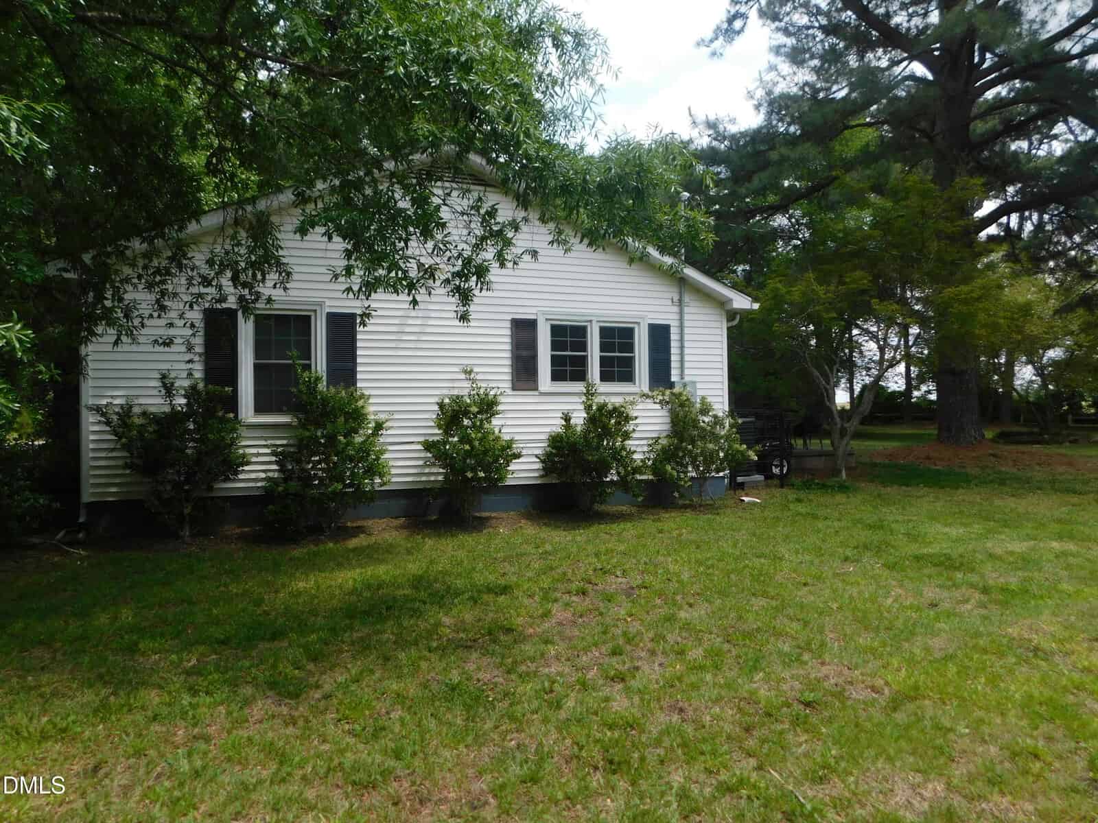A small white house with black shutters and several green bushes in front, set on a grassy yard with tall trees in the background under a partly cloudy sky.