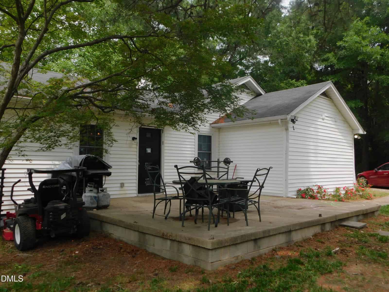 A white house with a gray roof, surrounded by trees. There is a concrete patio with a black metal table and chairs, and a grill placed nearby. A red vehicle is partially visible on the right.
