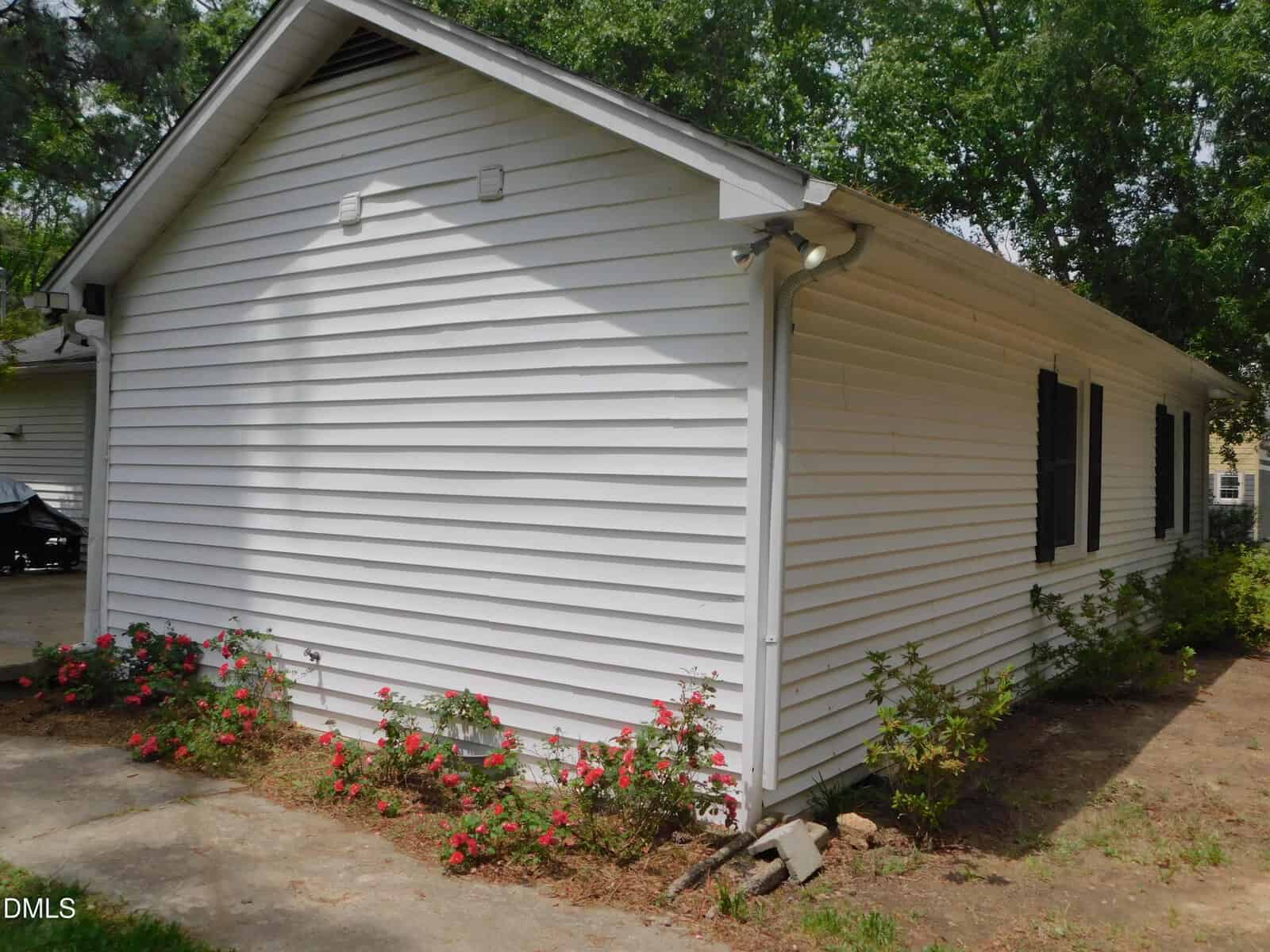 Side view of a white house with horizontal siding, black shutters on the windows, and a garden bed along the foundation with blooming red and pink flowers, next to a concrete walkway and surrounded by green trees.
