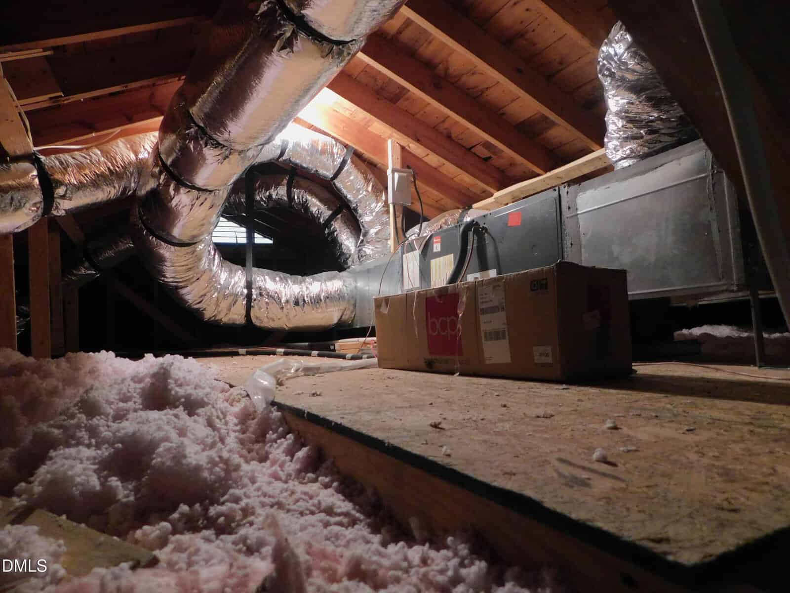 An attic with exposed wooden beams, ductwork wrapped in insulation, a large HVAC unit, cardboard boxes, and scattered pink fiberglass insulation on the plywood floor.