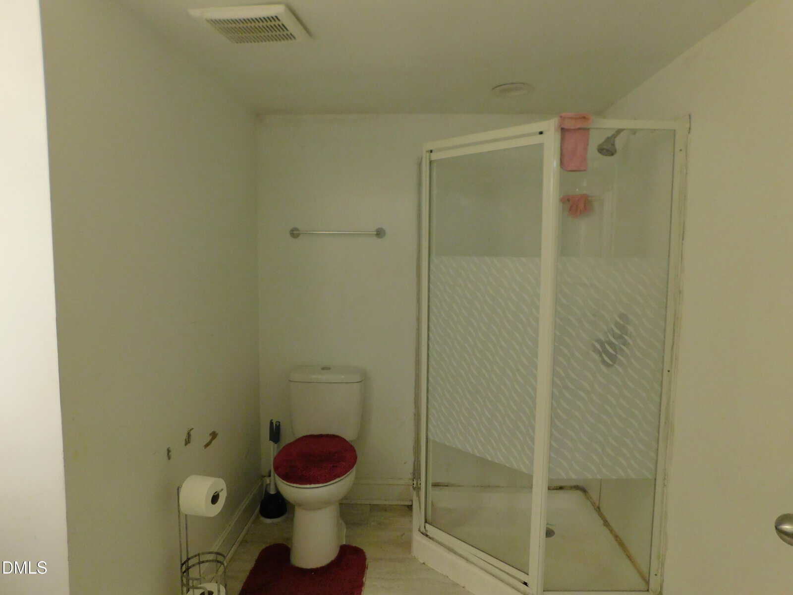 A small bathroom with a toilet and a glass shower stall, both with red mats. There is a towel rack on the wall and a toilet paper holder next to the toilet. The walls and floor are light-colored.