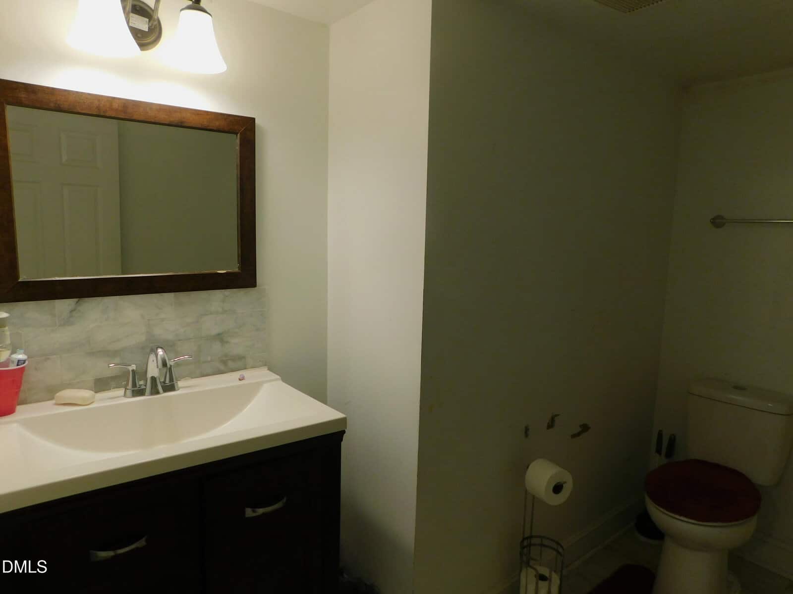 A bathroom with a sink, mirror, and light fixture on the left, and a toilet with a red seat cover on the right. There are some missing fixtures or marks on the wall next to the toilet.